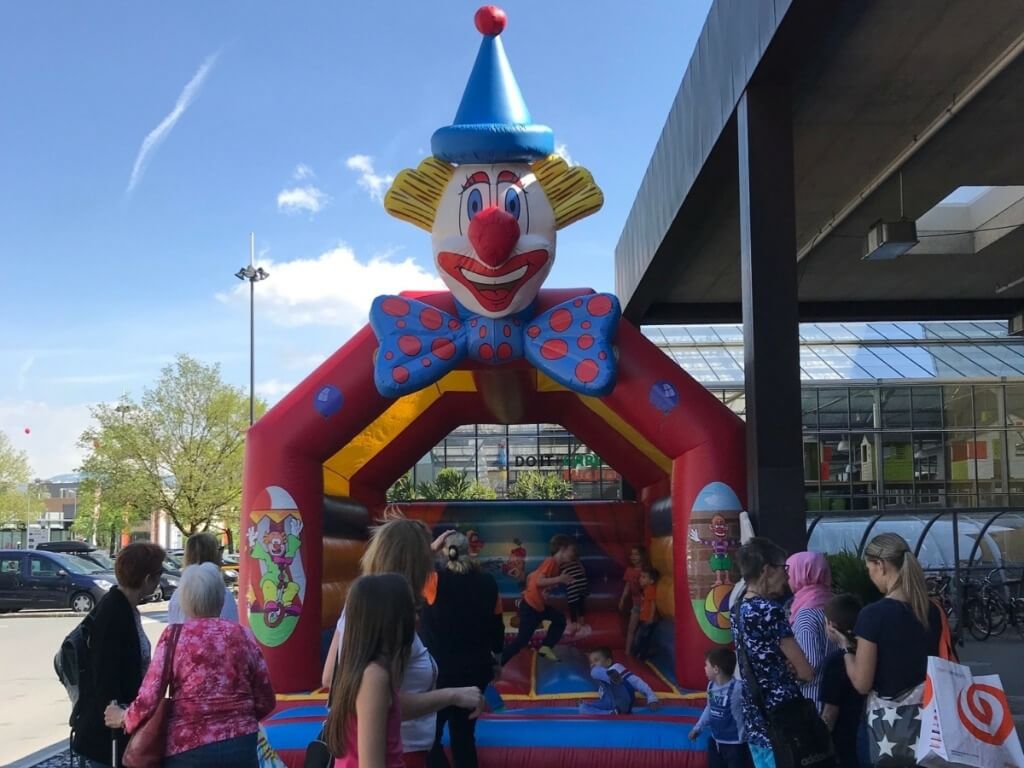 Kinder spielen auf der bunten 5 Meter Hüpfburg mit großem, wackelndem Clownskopf bei sonnigem Wetter.
