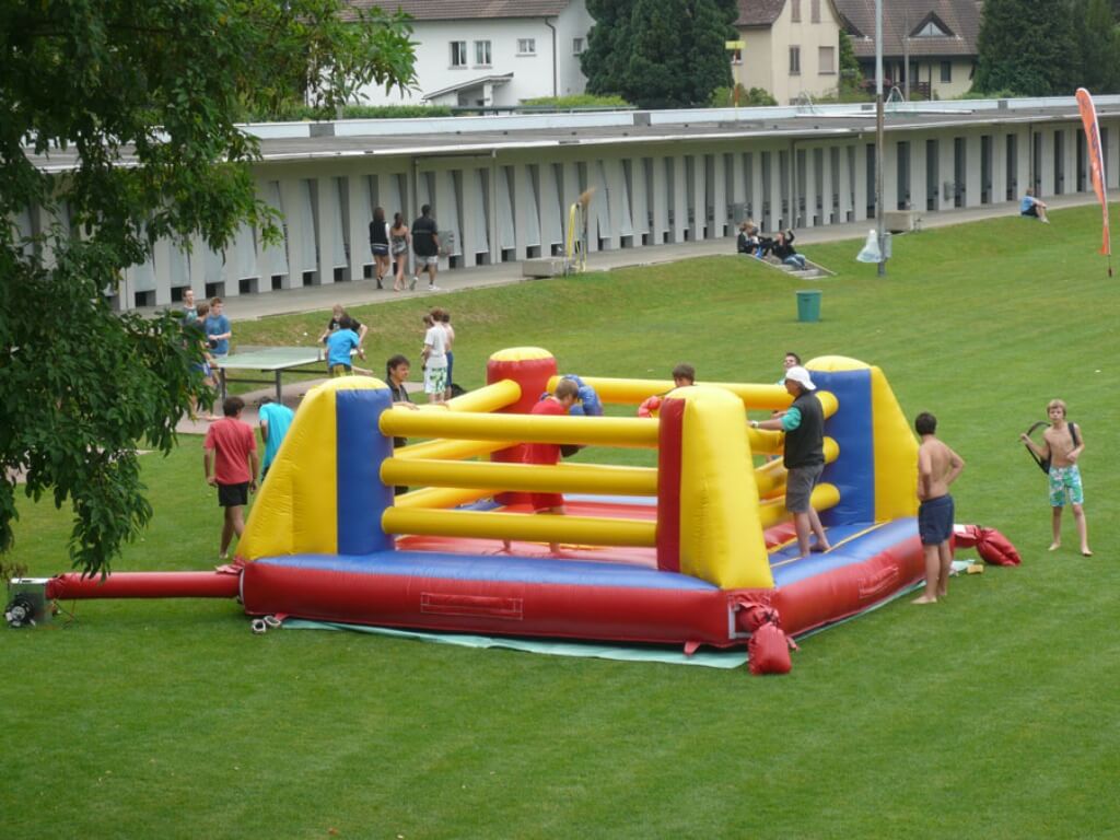 Zwei Personen kämpfen mit riesigen Boxhandschuhen in einem bunt aufgeblasenen Boxring auf einer Wiese.