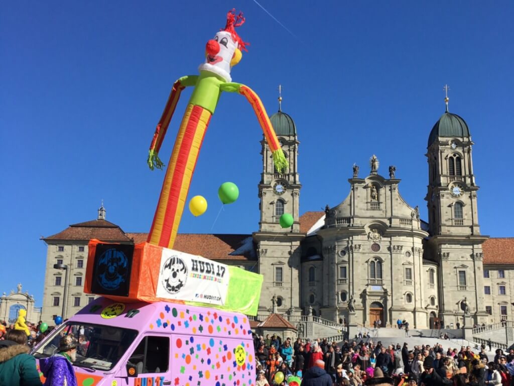 Großer bunter Skydancer mit lachendem Clownkopf auf einem bemalten Fahrzeug vor einer barocken Kirche bei blauem Himmel und vielen Besuchern.
