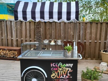 Mobiler Eistruhen-Wagen mit schwarzem Unterbau, buntem Schriftzug Ice Cream und gestreiftem Sonnendach