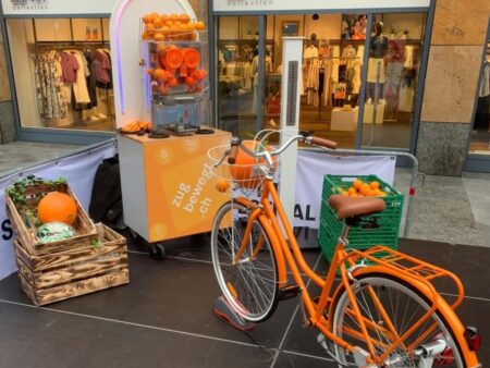 Orangefarbiges Fahrrad vor einer Saftpresse mit frischen Orangen in Kisten und Behältern auf einem Event-Stand.