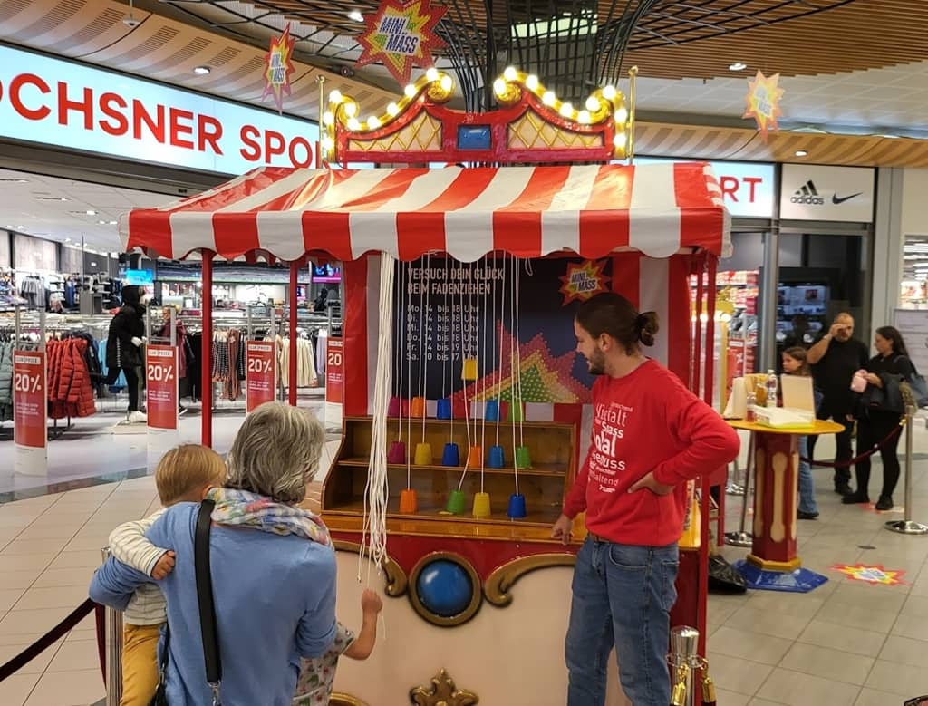 nostalgischer marktstand in einem einkaufszentrum. eine dame mit einem kleinkind auf dem arm versucht ihr glück beim glücksspiel fadenzeihen.