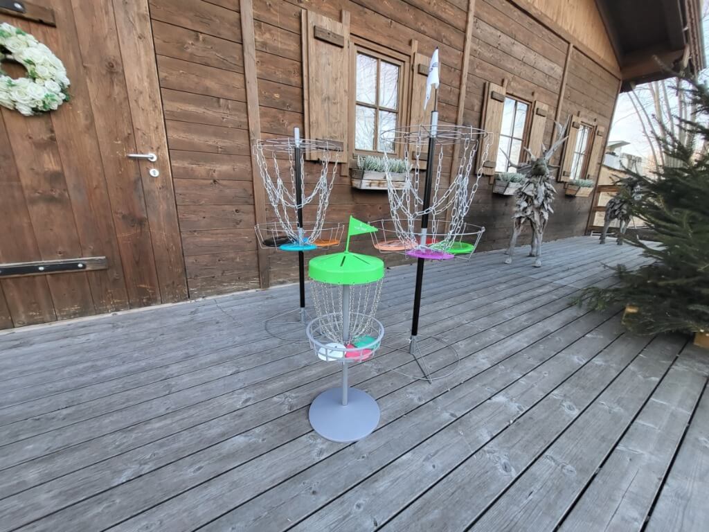 Drei Frisbee Golf Fangkörbe mit farbigen Discs stehen auf einer Holzterrasse vor einer rustikalen Holzhütte.