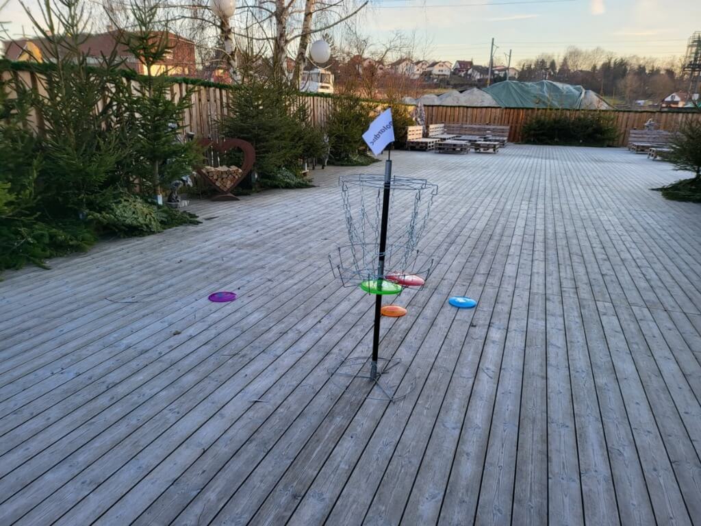 Tragbarer Frisbee Golf Fangkorb steht auf einer Holzterrasse, umgeben von bunten Frisbee-Scheiben.