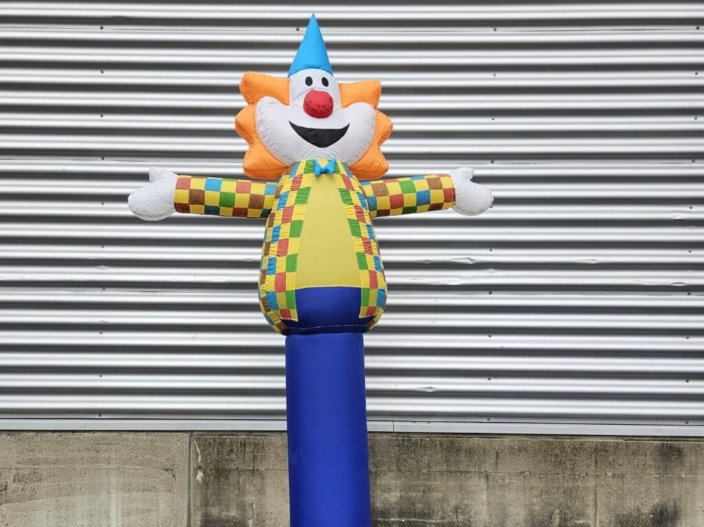 Bunter Skydancer Clown mit blauem Hut und ausgestreckten Armen vor Metallwand.