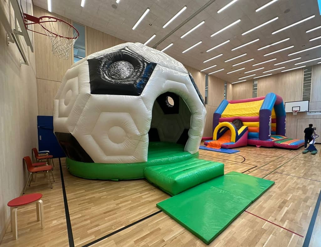 grosse runde hüpfburg in form eines fussballes. sie steht neben einer anderen bunten hüpfburg in einer turnhalle.