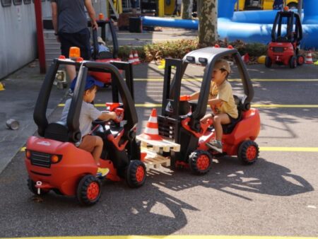 Zwei Kinder fahren spielerisch mit roten Tret-Gabelstaplern und transportieren eine kleine Palette.