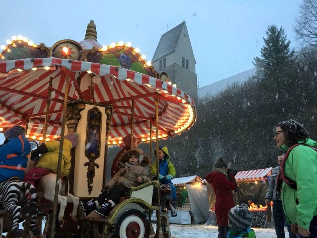 kinderkarussell auf einem weihnachtsmarkt im schneetreiben. die marktbesucher schauen zu wie die kinder auf dem nostalgiekarussell ihre runden drehen.