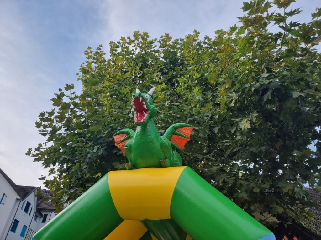 Fröhlicher grüner Drache mit orangen Flügeln als Dachfigur auf einer bunten Hüpfburg vor Baumkulisse.