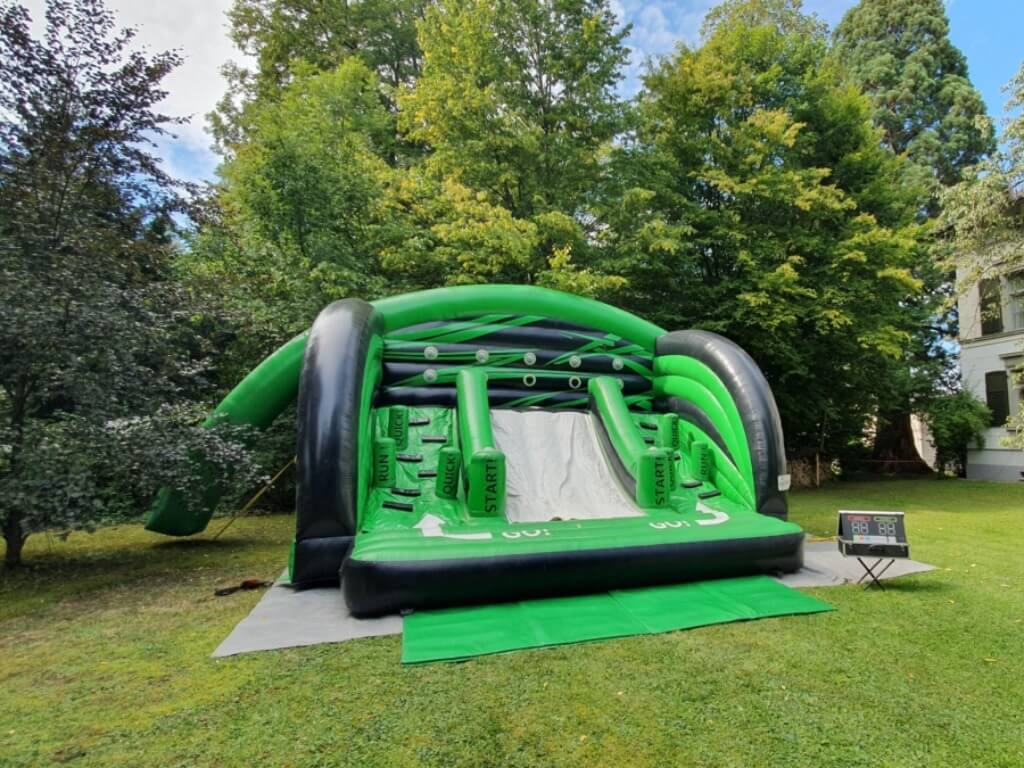 Grüner aufblasbarer Hindernisparcours mit Startpunkten und digitalem Scoreboard im Garten.
