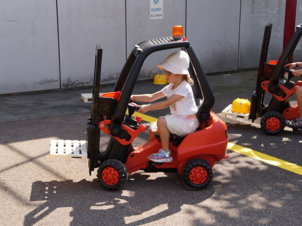 Kleines Mädchen steuert einen roten Kinder-Gabelstapler auf asphaltierter Fläche.