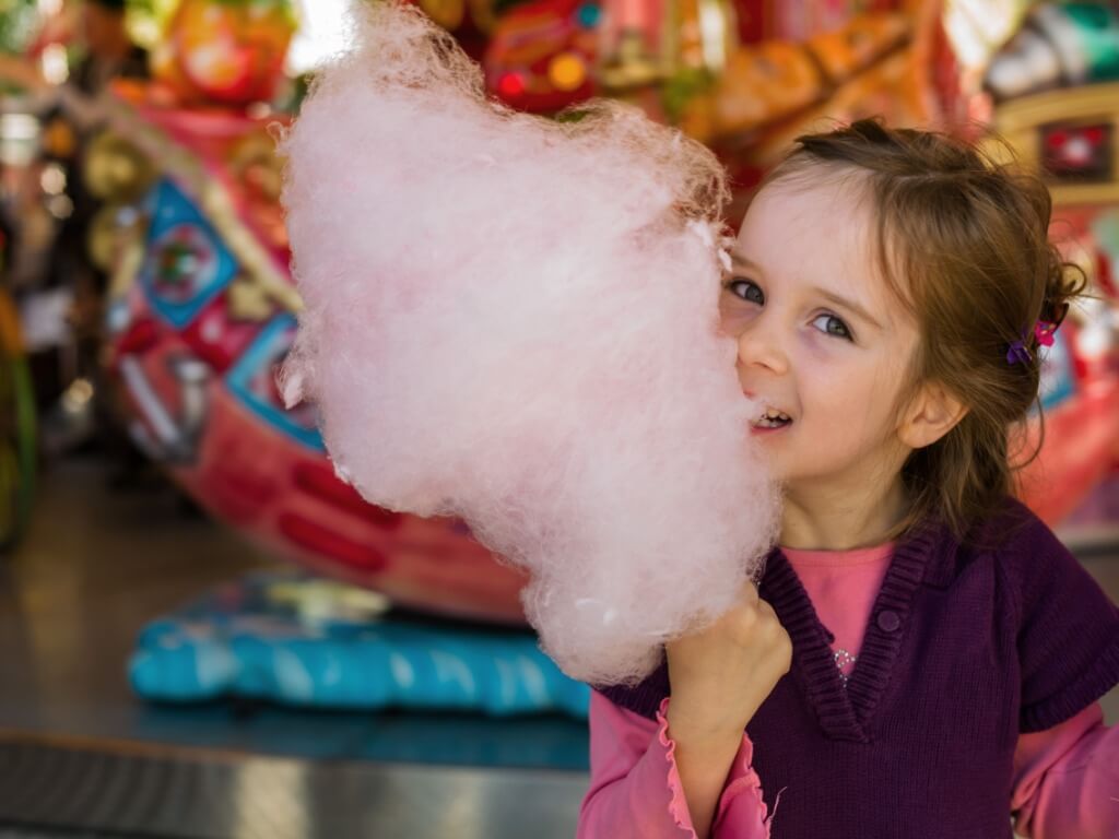 Ein Mädchen isst fröhlich eine große, rosa Zuckerwatte vor einem bunten Karussell.