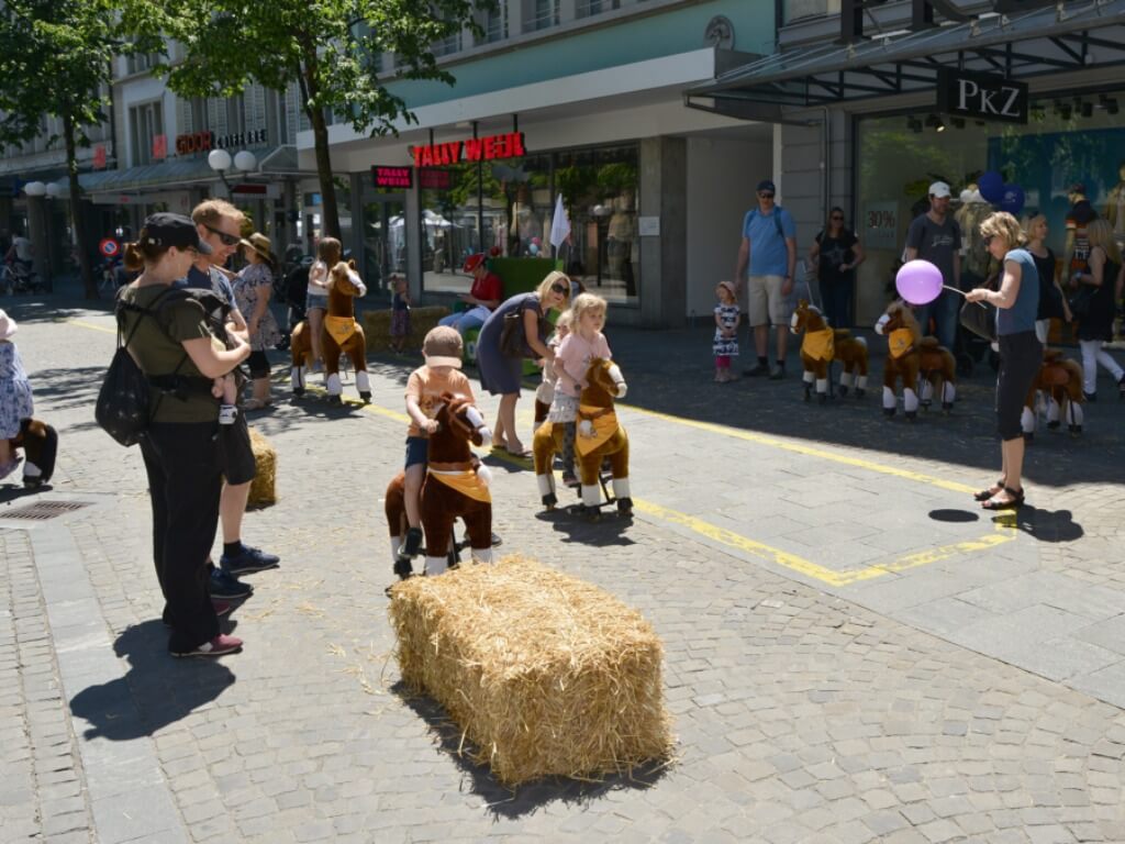Mehrere Kinder reiten auf Plüschpferden in einem Stadtparcours, umgeben von Erwachsenen und Publikum.