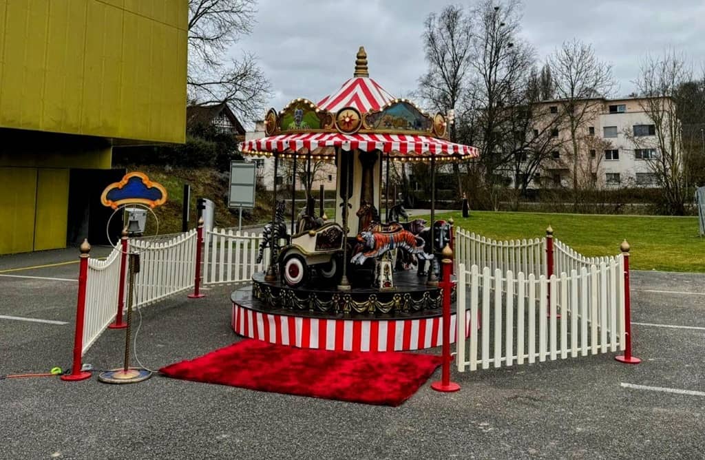 nostalgiekarussell in rot weiss. drumherum ein weisser zaun als dekoration mit roten pfosten. vor dem karussell liegt einaldend ein roter teppich.