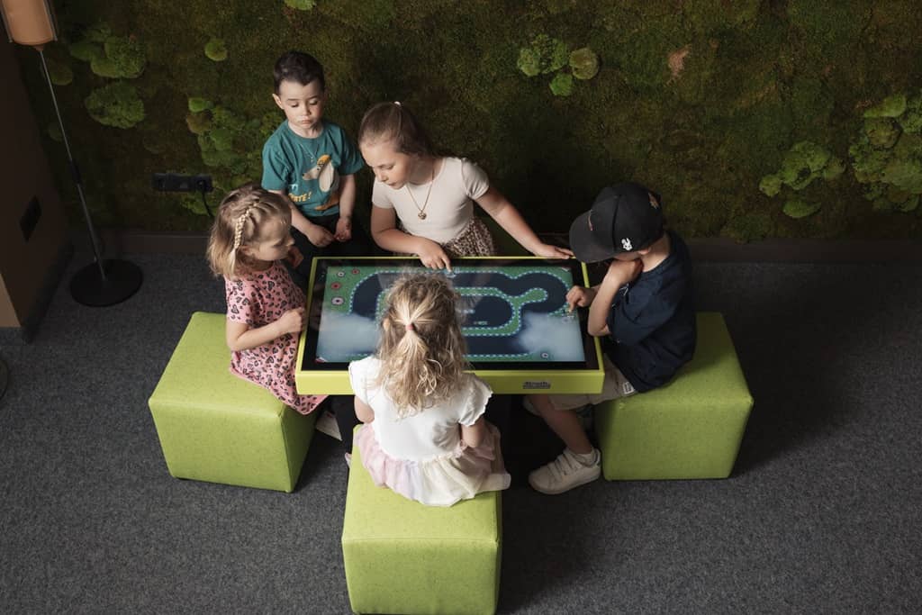 4touch spieletisch aus österreich. 4 kinder sitzen auf grünen hockern um diesen herum und haben spass beim spielen.