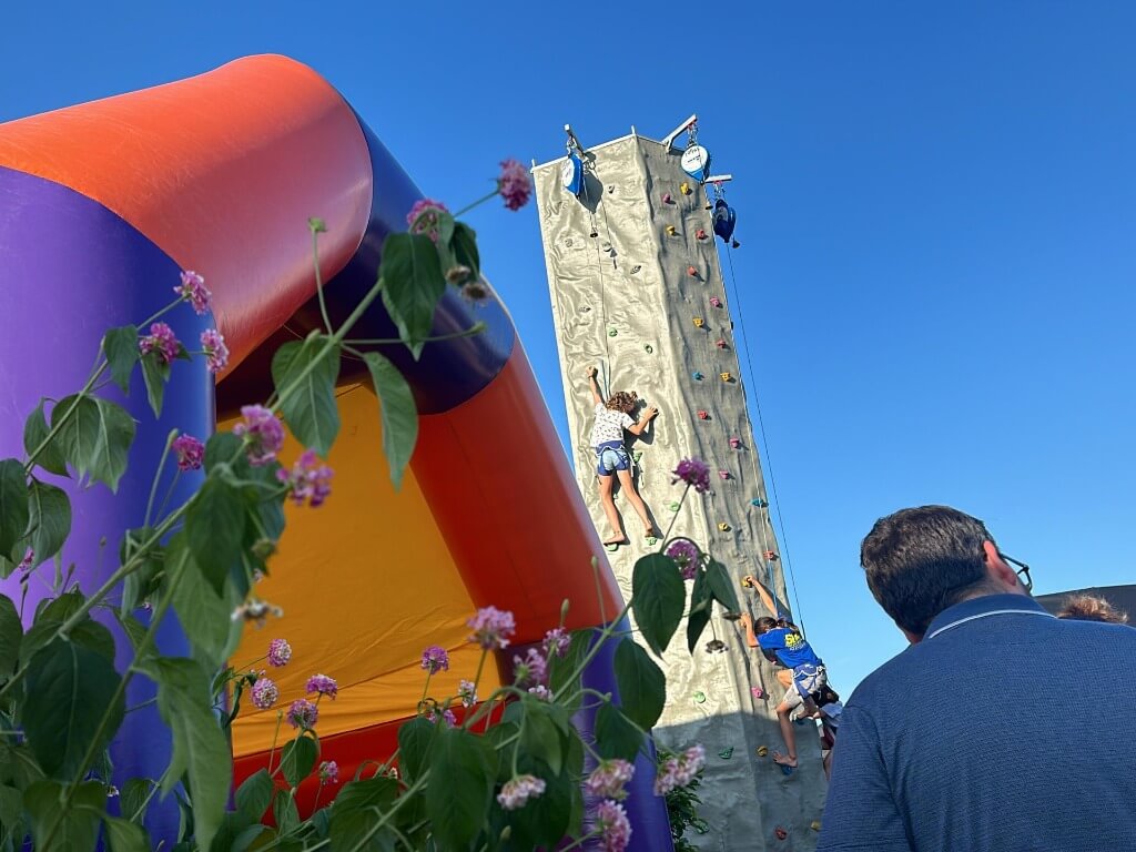 Kinder klettern gesichert an der acht Meter hohen mobilen Kletterwand The Rock bei sonnigem Himmel.