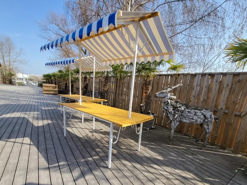 Marktstand mit blau-weiß gestreiftem Dach und gelber Arbeitsfläche auf Holzterrasse neben Holzlattenzaun und Skulptur