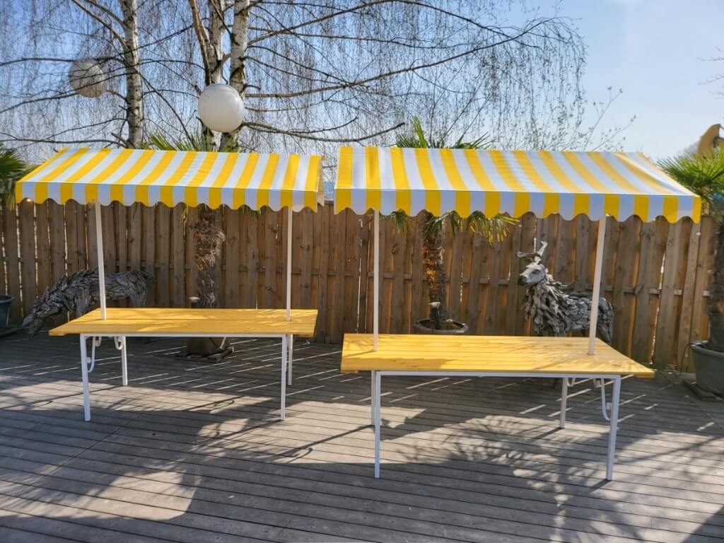 Zwei Marktstände mit gelb-weiss gestreiftem Dach und gelber Arbeitsfläche auf Holzterrasse vor Holzzaun.