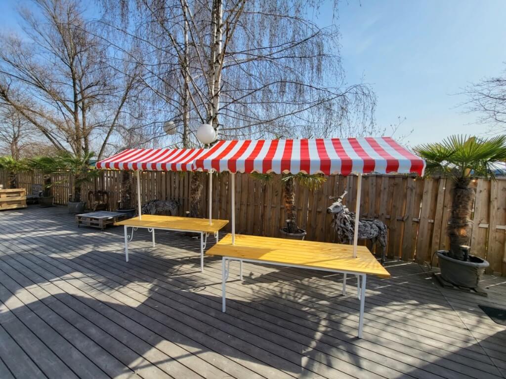 Zwei nebeneinander platzierte Marktstände mit rot-weissen Dächern auf einer Terrasse mit Palmen