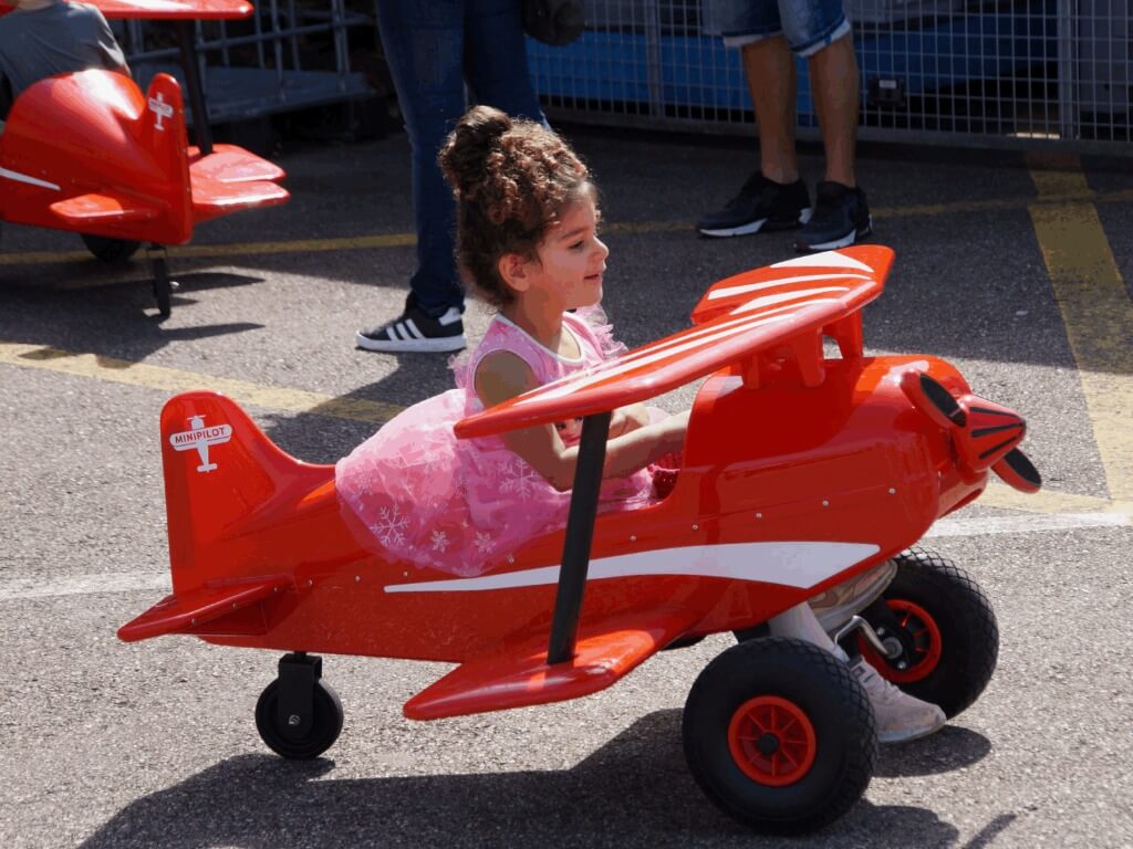 Ein Mädchen in rosa Kleid fährt glücklich ein rotes Mini Pilot Tretflugzeug auf Asphalt.