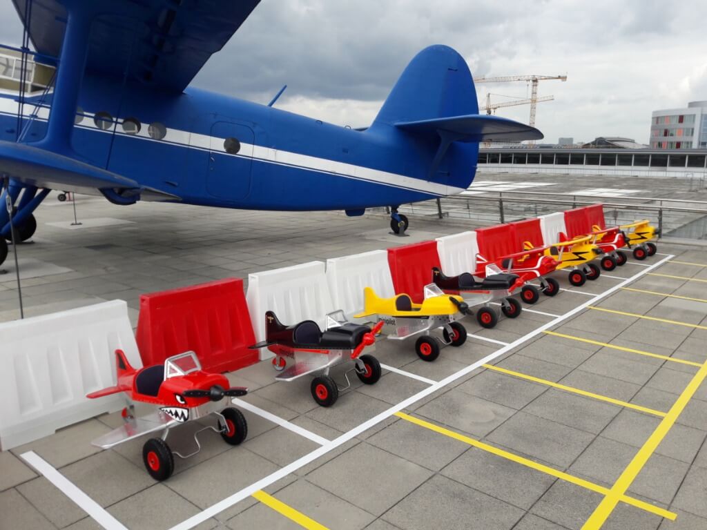 Reihe bunter Mini Pilot Tretflugzeuge auf einem Außenparkplatz neben einem großen blauen Flugzeug.