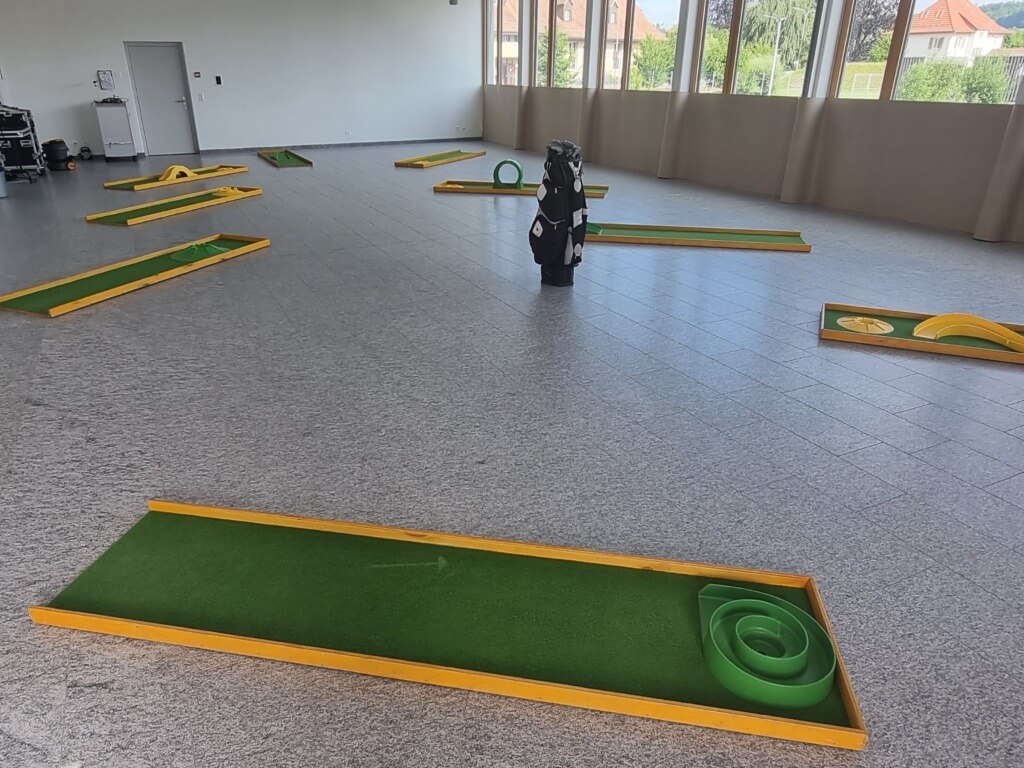 Neun Minigolfbahnen mit grünem Kunstrasen und gelben Umrandungen in einem großen, hellen Raum mit Fenstern und Golf-Equipment in der Mitte.