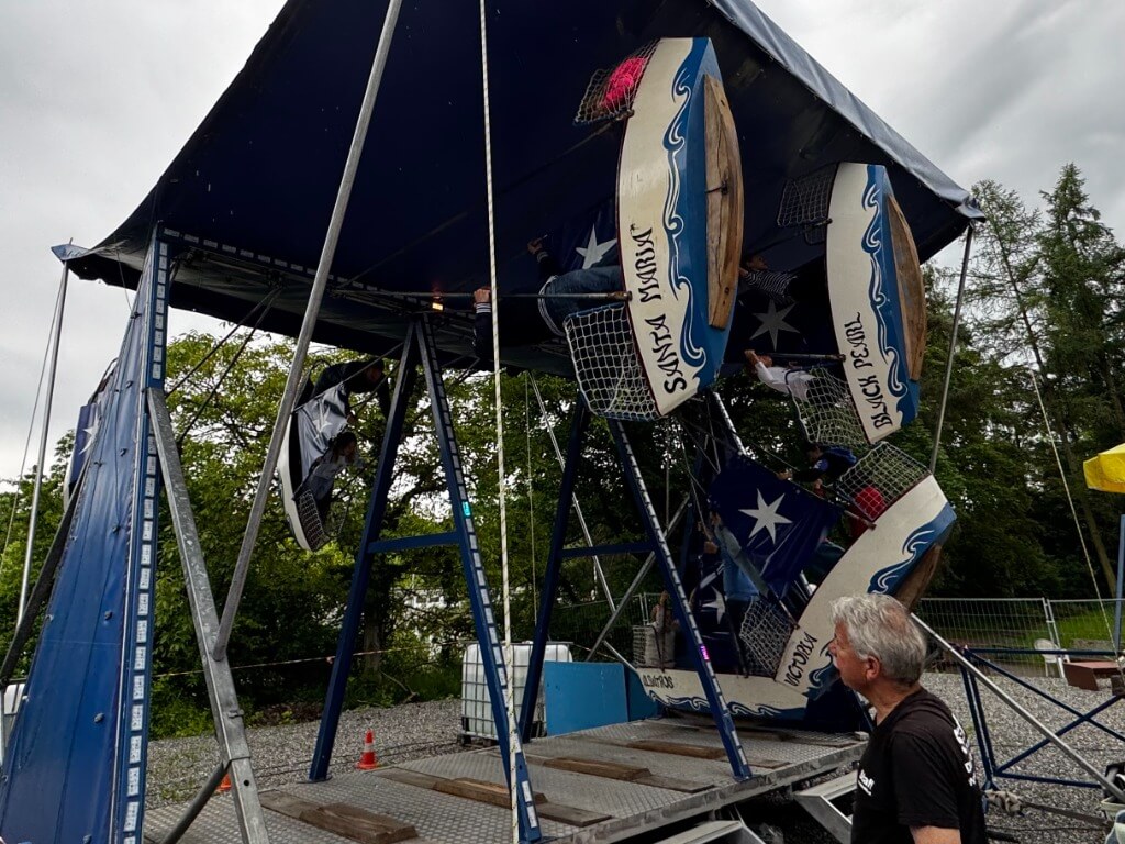 Schifflischaukel mit sechs Schiffen und Personen, teilweise unter einem blauen Zeltdach.