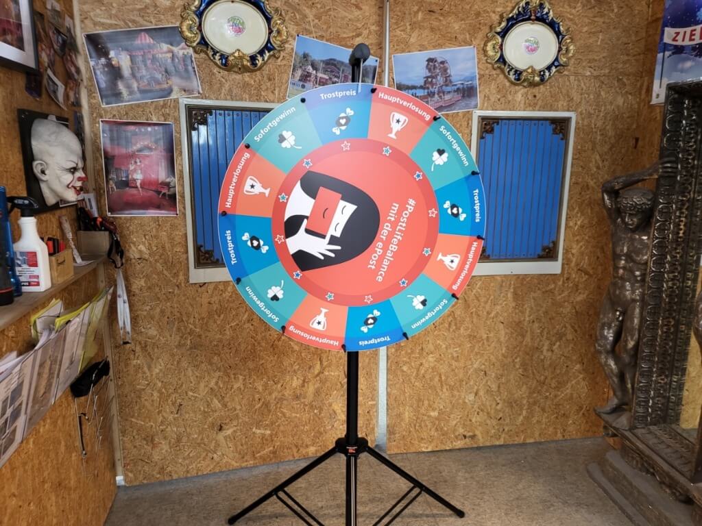 Glücksrad mit bunten Gewinnfeldern und individuellem Logo auf einem schwarzen Stativ in einem Holzraum mit Dekoration