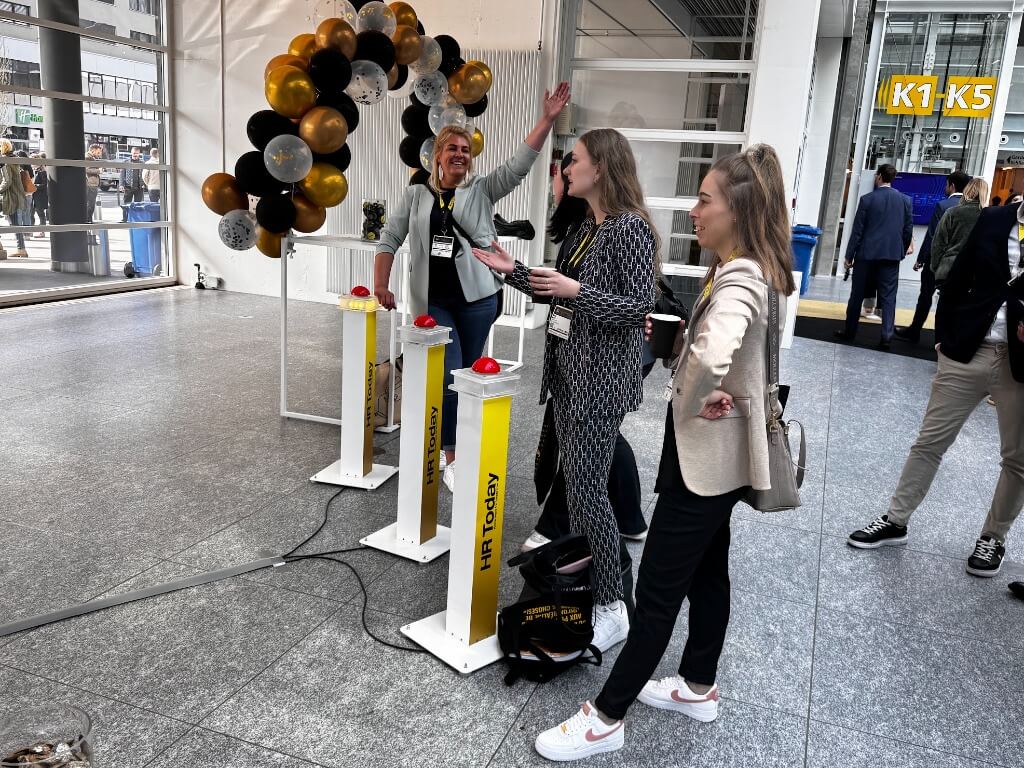 Drei Frauen stehen an einer Buzzer-Anlage mit leuchtenden roten Buzzern vor einem Ballonbogen in einem modernen Messegebäude.