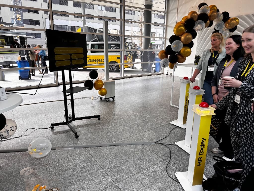 Drei Frauen an einem HR Today Buzzer-Pult bei einem interaktiven Quiz-Event in einem modernen Innenraum mit großen Fenstern.
