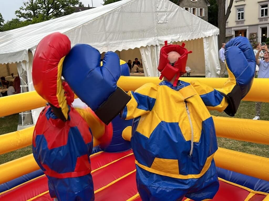 Zwei Personen in bunten aufblasbaren Boxanzügen mit großen Handschuhen in einem gelb-roten Boxring.