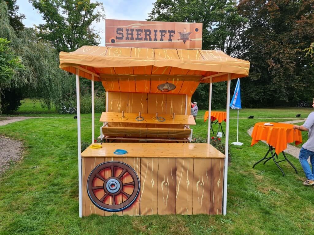 Spielstand zum Ringewerfen mit Western-Dekoration und Sheriff-Schild in einem grünen Park.