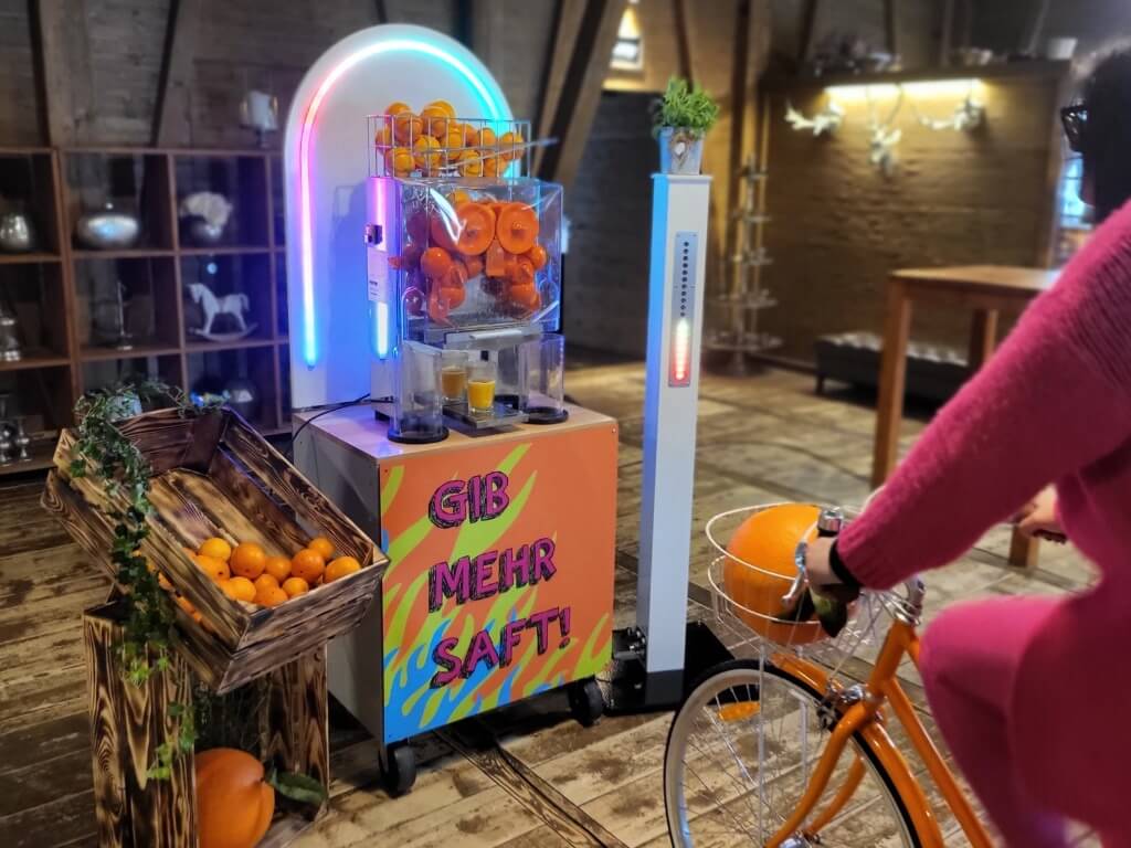 Person tritt in orangefarbenes Saftfahrrad und presst damit frischen Orangensaft an der Saftbar.