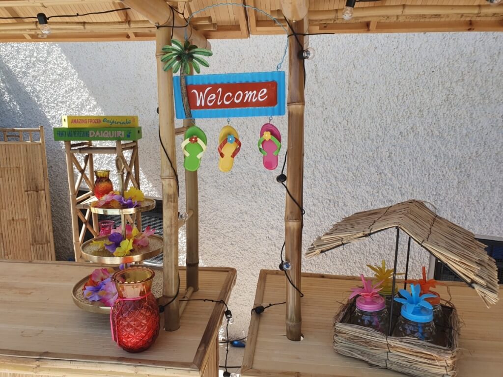 Bambus-Tiki-Bar mit Welcome-Schild, bunten Flip-Flop-Anhängern und dekorativen Gläsern mit Blumen