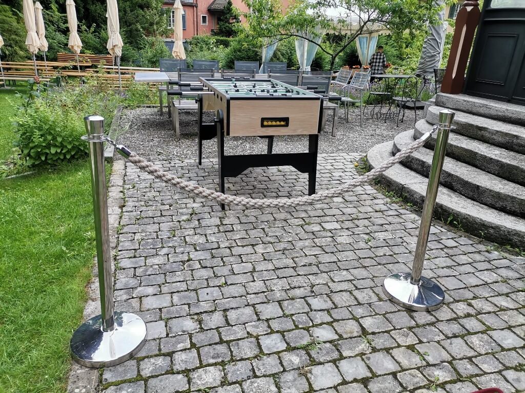 Moderner Töggelikasten steht auf gepflastertem Weg im Garten zwischen Absperrständern.