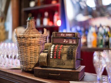 Alte, schwere Metallkasse mit rot-weißen und schwarzen Knöpfen steht neben einer geflochtenen Flasche auf einer Bar.