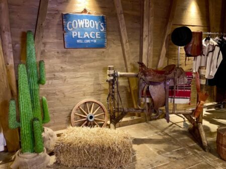Western-Dekoration mit blauem Holzschild ‚COWBOY'S PLACE‘, Sattel auf Holzständer, Heuballen und Deko-Kaktus in rustikalem Holzzelt.
