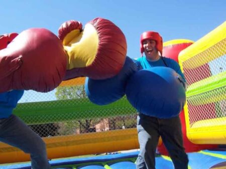 Zwei Personen mit überdimensionalen aufblasbaren Boxhandschuhen kämpfen im farbenfrohen aufblasbaren XXL Boxring unter blauem Himmel.