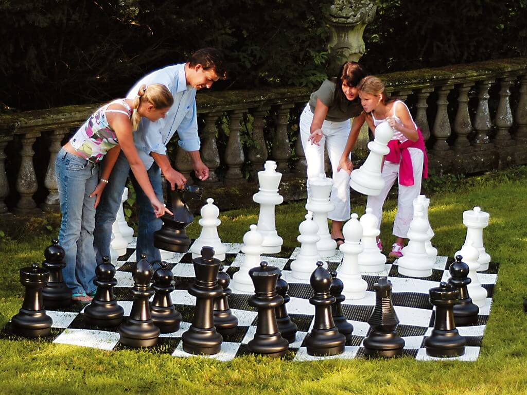 Vier Personen spielen zusammen ein lebensgroßes Gartenschachspiel auf einer Wiese.