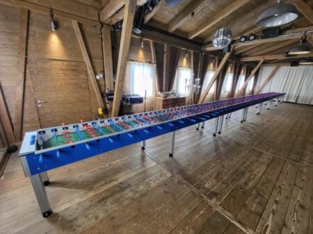 Extrem langer Tischfussballtisch mit Blauton und zahlreichen roten und blauen Spielfiguren, aufgestellt in einem hellen Holzbodenraum.